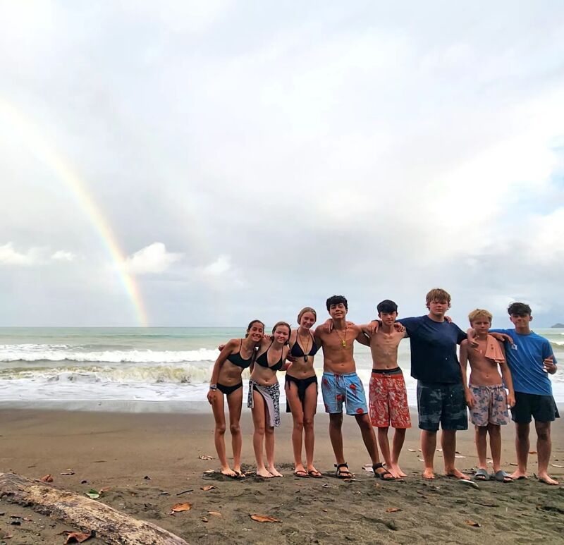 A group of eight young people stand on a sandy beach with a rainbow arcing over the ocean in the background. They are dressed in swimwear and casual beach attire, some with their arms around each other. The sky is overcast, but the scene is brightened by the rainbow and the smiles of the group. The waves are gently rolling onto the shore.